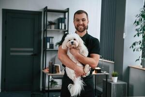 Man is smiling.. Groomer, is with, Maltese dog indoors. Man is in the black uniform photo