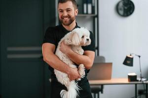 Man is, smiling. Groomer is with Maltese dog indoors. Man is in the black uniform photo