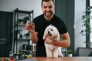 Smiling and holding scissors. Groomer is with Maltese dog indoors. Man is in the black uniform photo