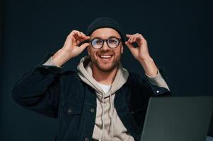 Smiling and, sitting by laptop. Handsome man is in the studio against background photo