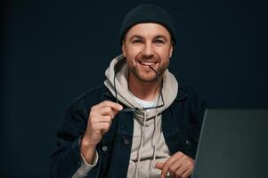 Smiling and sitting by laptop. Handsome man is in the studio against background photo