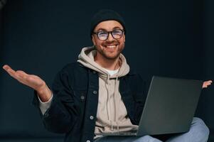 Smiling, sitting and using laptop. Handsome man is in the studio against background photo