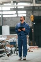 With tablet in hands. Factory ,worker in blue uniform is indoors photo