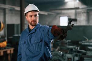 Metal wrench in hand. Factory worker in blue uniform is indoors photo