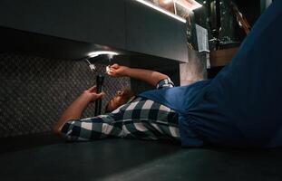 Using flashlight.. Fixing water tap. Plumber in blue uniform is at work in the bathroom photo