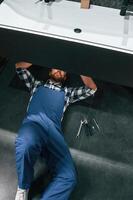 Lying down, on the floor, fixing the siphon. Plumber in blue uniform is at work in the bathroom photo