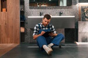 Sitting on, the floor with notepad in hands. Plumber in blue uniform is at work in the bathroom photo