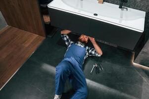 Lying ,down on the floor, fixing the siphon. Plumber in blue uniform is at work in the bathroom photo