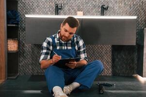 Sitting ,on the floor with notepad in hands. Plumber in blue uniform is at work in the bathroom photo