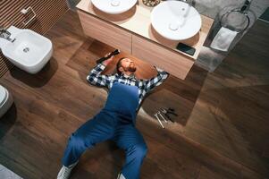 Top view. ..Lying down on the ground, fixing water tap. Plumber in blue uniform is at work in the bathroom photo