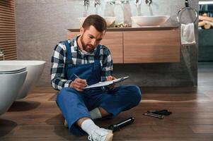 With notepad in hands. Plumber in blue uniform is at work in the bathroom photo
