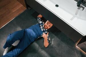 Lying down on the floor, fixing the siphon.. Plumber in blue uniform is at work in the bathroom photo