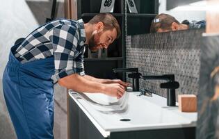 Side view.. Fixing wate,r tap. Plumber in blue uniform is at work in the bathroom photo