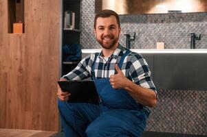 Sitting on the floor with notepad in hands. Plumber, in blue uniform is at work in the bathroom photo