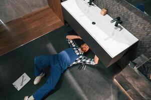 Fixing t,he siphone. Lying down on the floor. Plumber in blue uniform is at work in the bathroom photo