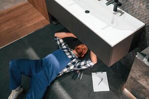 Fixing the siphone.. Lying down on the floor. Plumber in blue uniform is at work in the bathroom photo