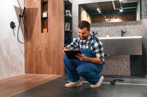 Sitting on the floor with notepad in hands. Plumber in blue uniform is at work in the bathroom photo