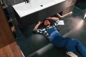 Fixing, the siphone.. Lying down on the floor. Plumber in blue uniform is at work in the bathroom photo