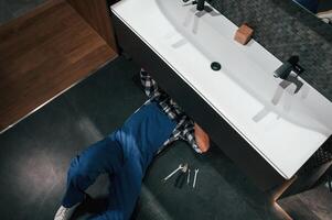 Lying down on the floor, fixing the siphon. Plumber in blue uniform is at work in the bathroom photo