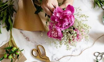 The photo showcases the art of floristry, with hands carefully positioning pink peonies on a piece of craft paper.