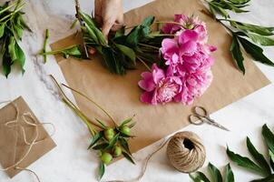 A floral composition in progress, featuring pink peonies, craft paper, scissors, and twine, suggesting a handmade and heartfelt creation. photo