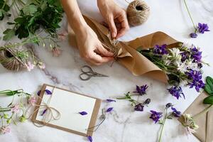 Delicate purple and white flowers are being carefully wrapped in brown paper, accompanied by a blank card and crafting tools. photo