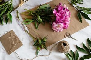 A close-up view of hands delicately handling pink peonies on craft paper, with tools and materials scattered around. photo