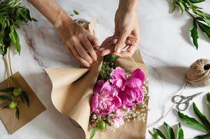 Hands carefully tying twine around a bouquet of pink peonies wrapped in brown paper on a marble surface. photo