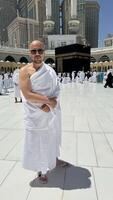 A bald man poses for a photo in front of the Kaaba at noon after performing Umrah at Masjid al-Haram, Mecca. A sacred Islamic pilgrimage in the heart of the holy mosque under the bright sunlight.