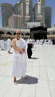 A bald man poses for a photo in front of the Kaaba at noon after performing Umrah at Masjid al-Haram, Mecca. A sacred Islamic pilgrimage in the heart of the holy mosque under the bright sunlight.