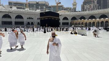 A bald man poses for a photo in front of the Kaaba at noon after performing Umrah at Masjid al-Haram, Mecca. A sacred Islamic pilgrimage in the heart of the holy mosque under the bright sunlight.