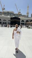 A bald man poses for a photo in front of the Kaaba at noon after performing Umrah at Masjid al-Haram, Mecca. A sacred Islamic pilgrimage in the heart of the holy mosque under the bright sunlight.