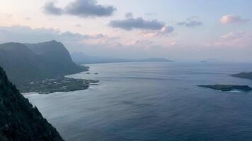 Aerial coastal view of Hawaii's serene ocean horizon during sunset, with pastel clouds, rugged mountains, and scattered islands completing the tranquil scene video