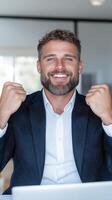 Satisfied businessman in a suit exults in front of a laptop in a modern office. He expresses joy and success by clenching his fists in a victory gesture. photo