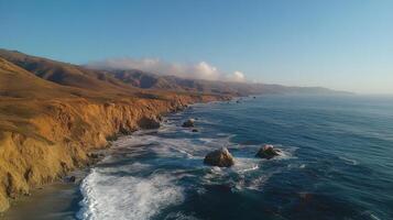 Ocean waves crash against rocky cliffs and beaches under a clear blue sky. Coastal hills and mountains rise in the background, shrouded by low clouds. photo