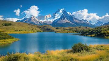 Mountain lake landscape. Blue water in foreground, grass and bushes, tall snowy peaks on the background. Sunny day with white clouds. photo