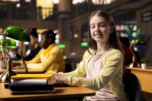 Portrait of a cute schoolgirl working on a project in the school library, using the academic database for homework research. Preteen student solving her school assignments for higher grades. photo