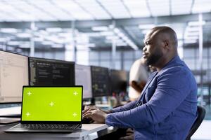 Server room programmer using green screen laptop to find firewall misconfigurations. African american man checking systems bottlenecks leading to sluggish data transfer rates using mockup notebook photo