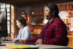 Call center employee using audio headset to answer customers technical inquiries about software issues. African american woman doing customer service job using PC to troubleshoot problems photo