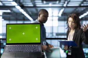 Green screen laptop in front of data center technicians in blurry background calibrating energy usage, inspecting UPS configurations, supporting sustainable infrastructure photo