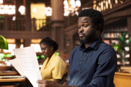 Black university student feeling anxious and struggling with learning, trying to memorize relevant information from class notes and research papers at the library. Male pupil studying to pass exams. photo