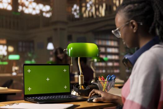 University student collecting notes from academic sources next to green screen, using the library database and books collection for scientific research. Young adult working on bachelor paper. photo