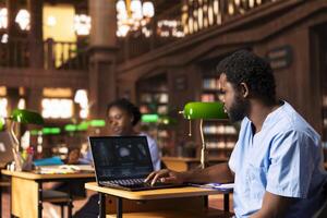 Black med school scholar studying and taking notes using online information, reviews a thesis project in the campus library. Young adult collecting academic data for medical research. photo
