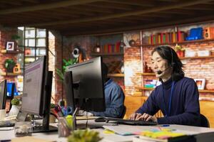 Senior call center agent in office helps customers by answering inquiries, addressing issues. Experienced Asian man in customer support workspace in front of PC using headset mic to talk with clients photo