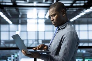 African american engineer in data center with laptop automating routine maintenance tasks, executing diagnostic scripts. Man adjusting cooling and power settings, validating server configurations photo