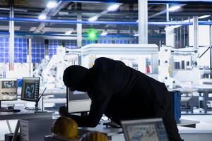 Masked hacker sneaking into solar panel plant at night, inserting USB stick into PC to steal confidential manufacturing data. Thief in photovoltaics facility extracts company files and uploading malware photo