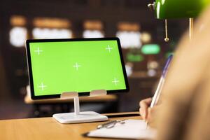 College student writes her homework next to isolated chroma key screen on tablet, using various books at the campus library to finish her project for a high grade. Academic resources for research. photo