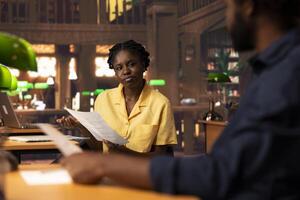 African american team comparing class notes and doing research at the library, consulting the academic database for exam preparation. Two pupils doing teamwork for educational purposes. photo