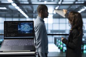 Laptop in front of data center engineers in blurry background deploying artificial intelligence tools. Close up of AI coding on notebook screen next to technicians inspecting server clusters photo