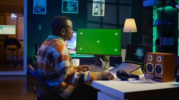 Black person programming neural network code with a green screen on computer to build databases. System engineer working from home while looking at data center security servers. Camera B. photo